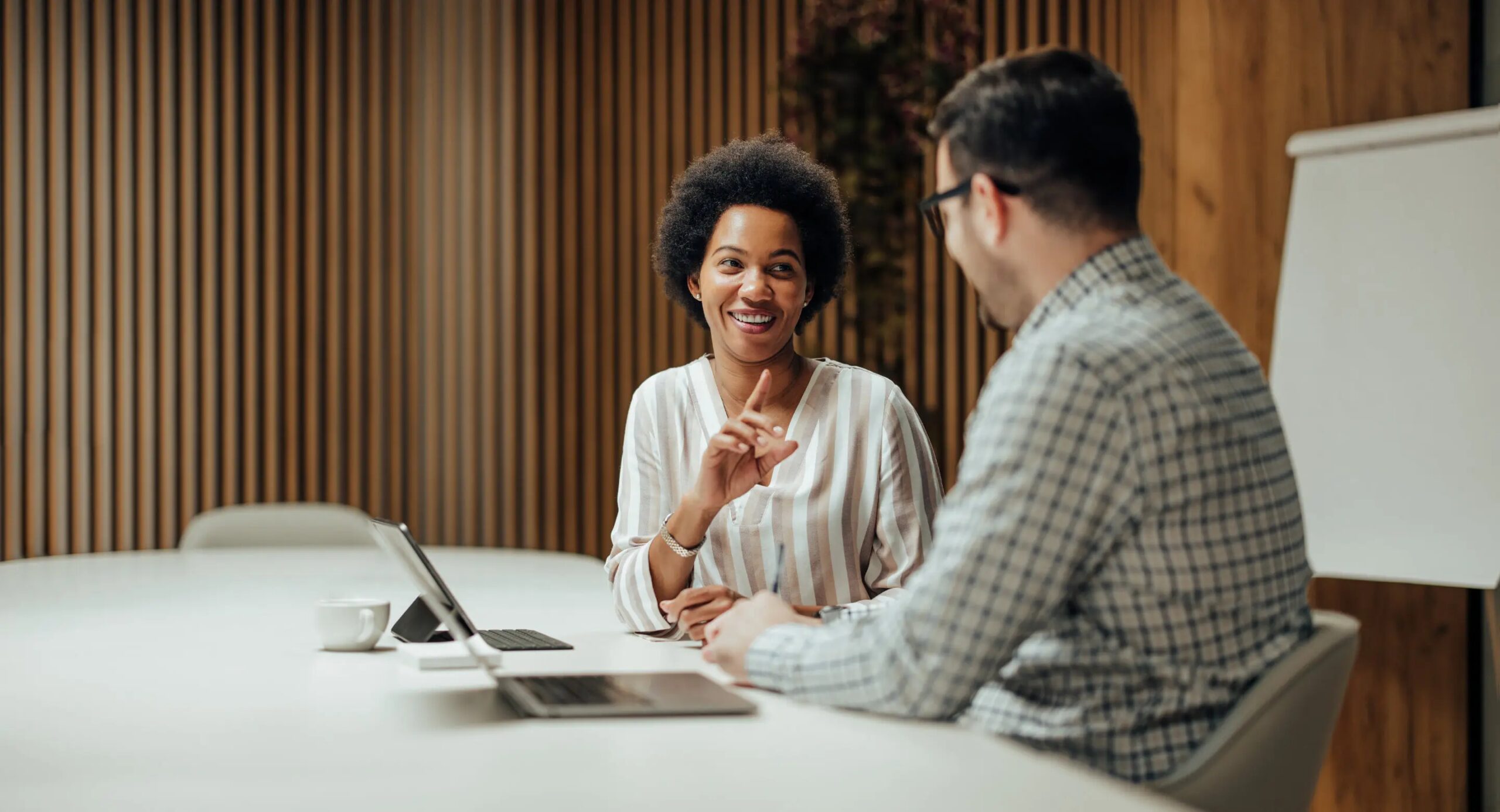 Two people talking in an office with wood panel background, one woman smiling and a man with glasses in checkered shirt