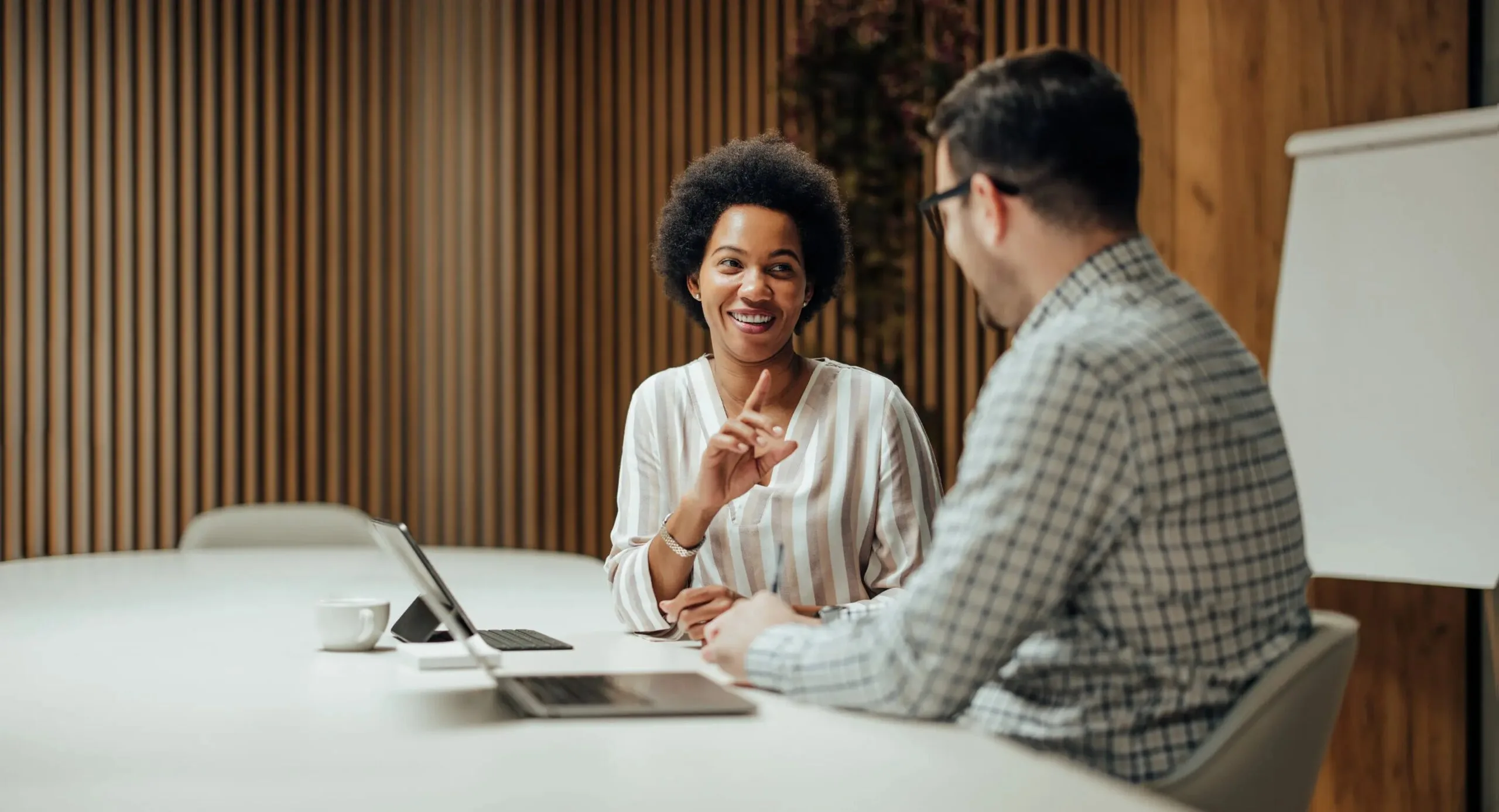 Two people talking in an office with wood panel background, one woman smiling and a man with glasses in checkered shirt