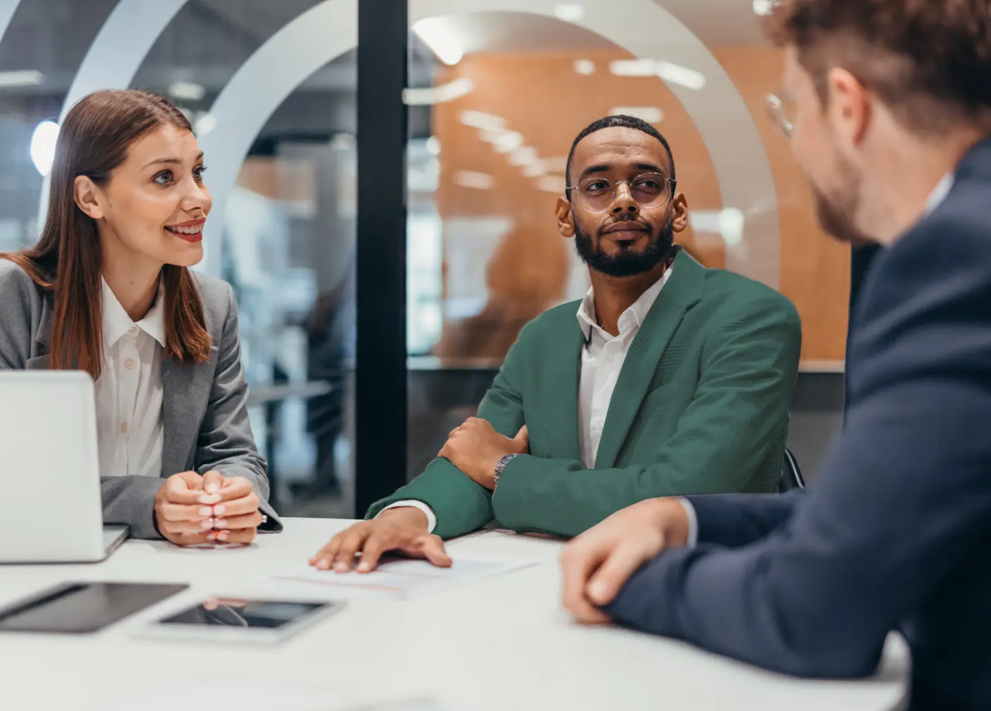 Three business professionals in a meeting room, one woman and two men, discussing with smiles and attentive expressions