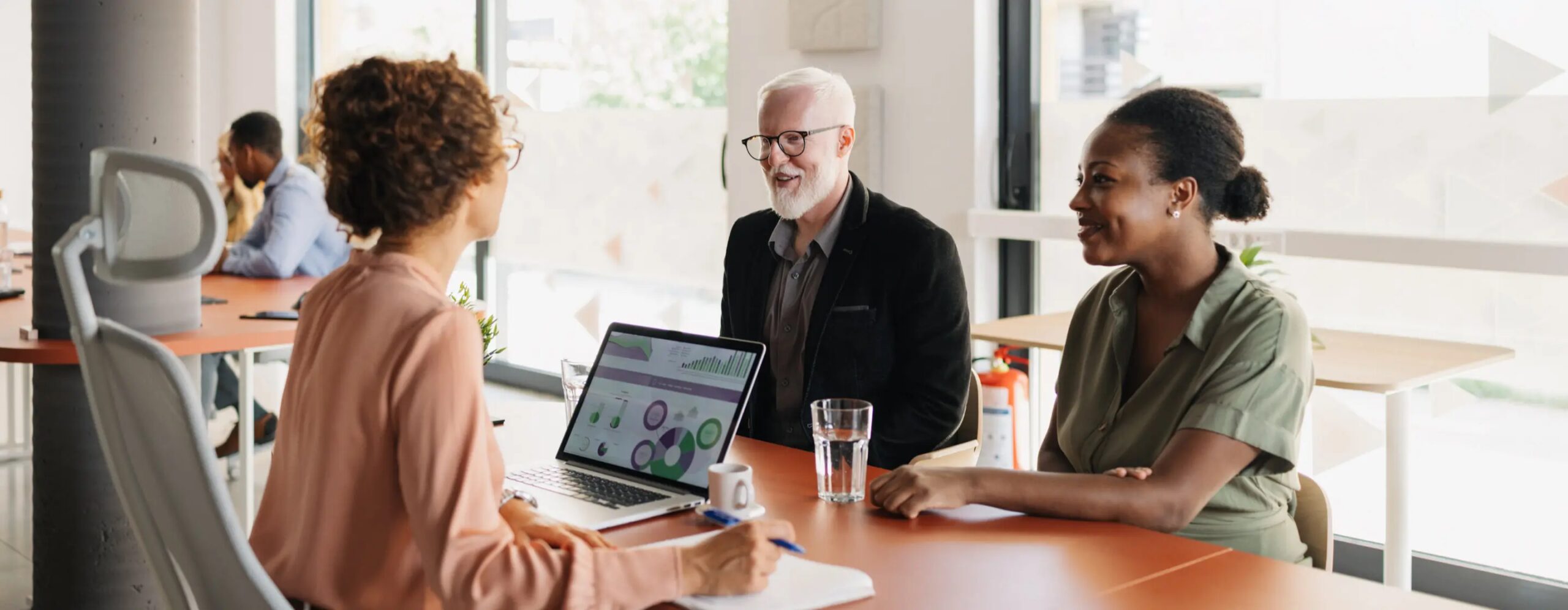 Office meeting scene with three people sitting around a table, one person with a laptop showing charts
