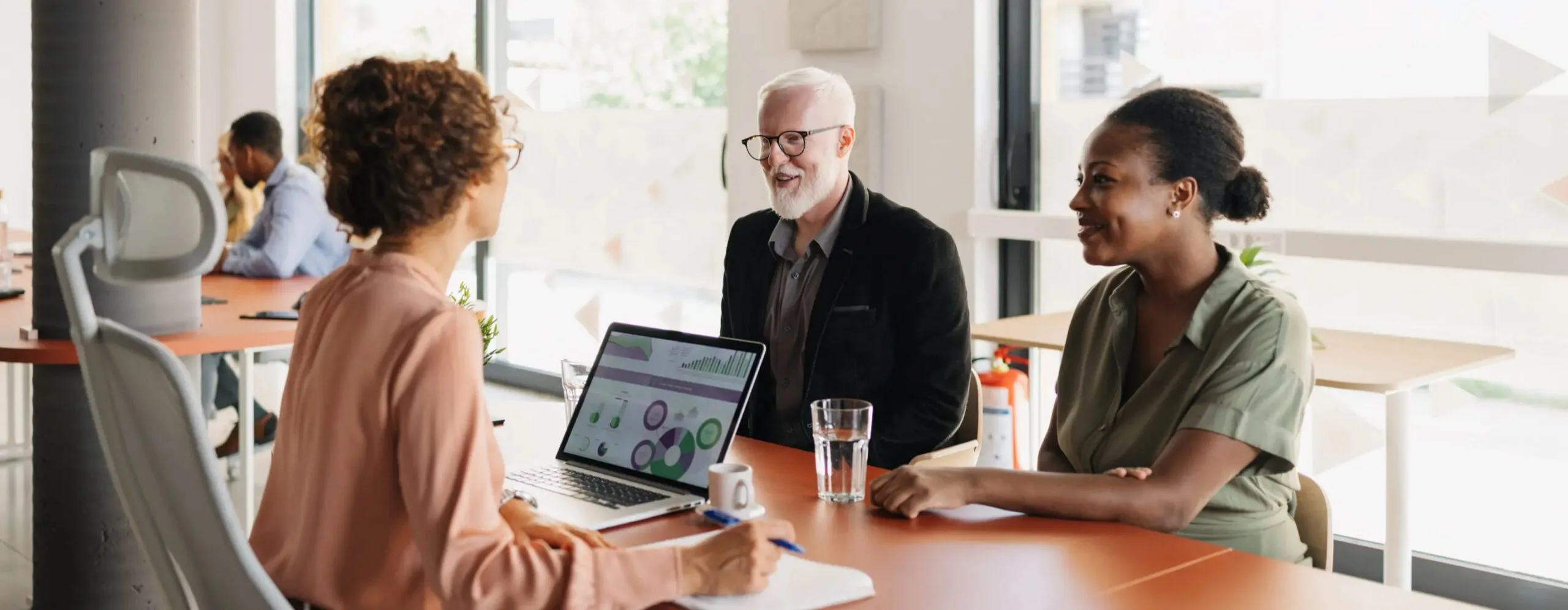 Three people in a bright modern office meeting, one with a laptop showing charts
